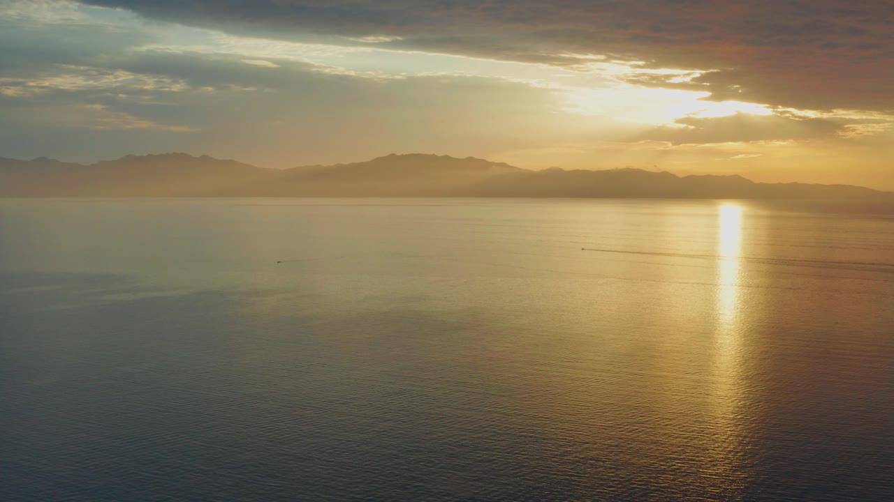 Wide aerial view over open water in El Sargento La Ventana, Mexico, with golden sun breaking through clouds, distant mountains on the horizon, and no visible people.