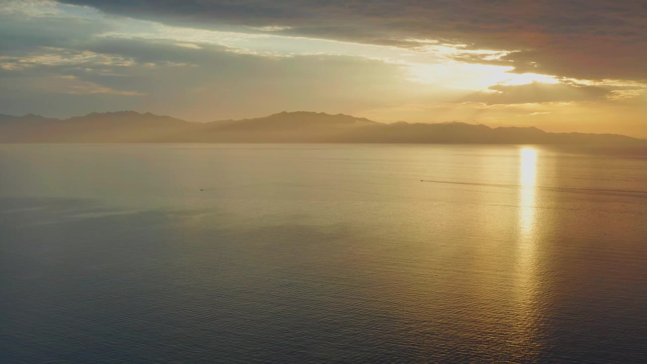 Aerial coastal view in El Sargento La Ventana, Mexico, showing a bright vertical sun reflection on the sea, layered clouds above, distant mountains, and no visible people.
