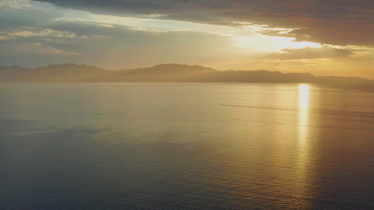 A high view of the El Sargento La Ventana coastline area shows the sun reflection stretching down the water, dark mountains across the background, cloud bands overhead, and no visible people.