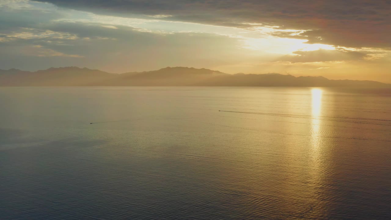 Aerial ocean sunset with a bright vertical reflection and small wake lines.