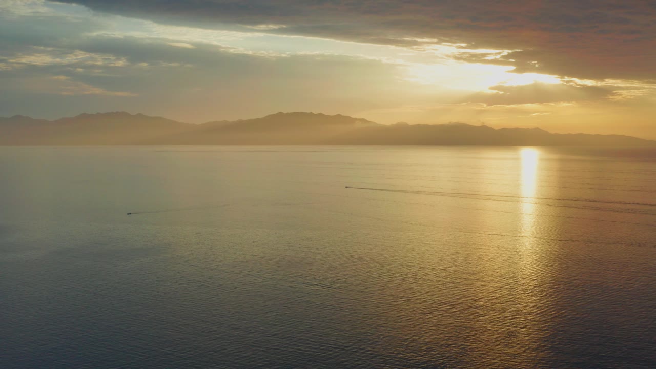 Opening frame of sunrise over the sea at El Sargento La Ventana, Mexico, with a bright reflection on the water, distant mountain silhouettes, cloud cover, and no visible people.
