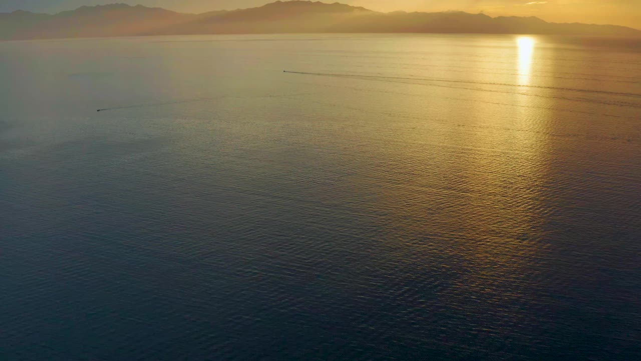 High view over the sea in El Sargento La Ventana, Mexico, with the sun glare bright on the right, layered mountains along the horizon, faint wake trails crossing the midwater, and no visible people.