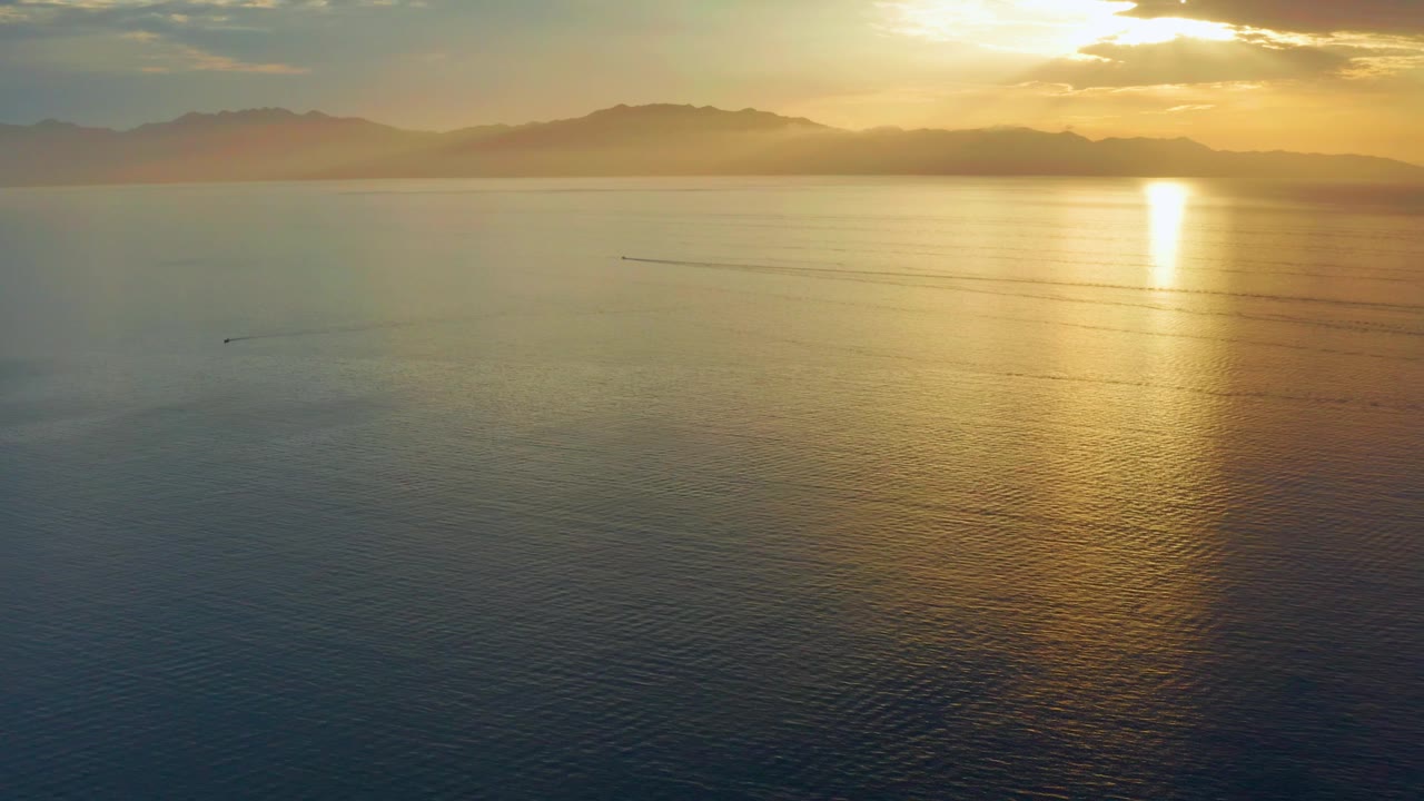 Mid-clip view over the sea in El Sargento La Ventana, Mexico, with a softer gold reflection on the right, hazy mountain shapes across the horizon, small wake lines in the center, and no visible people.