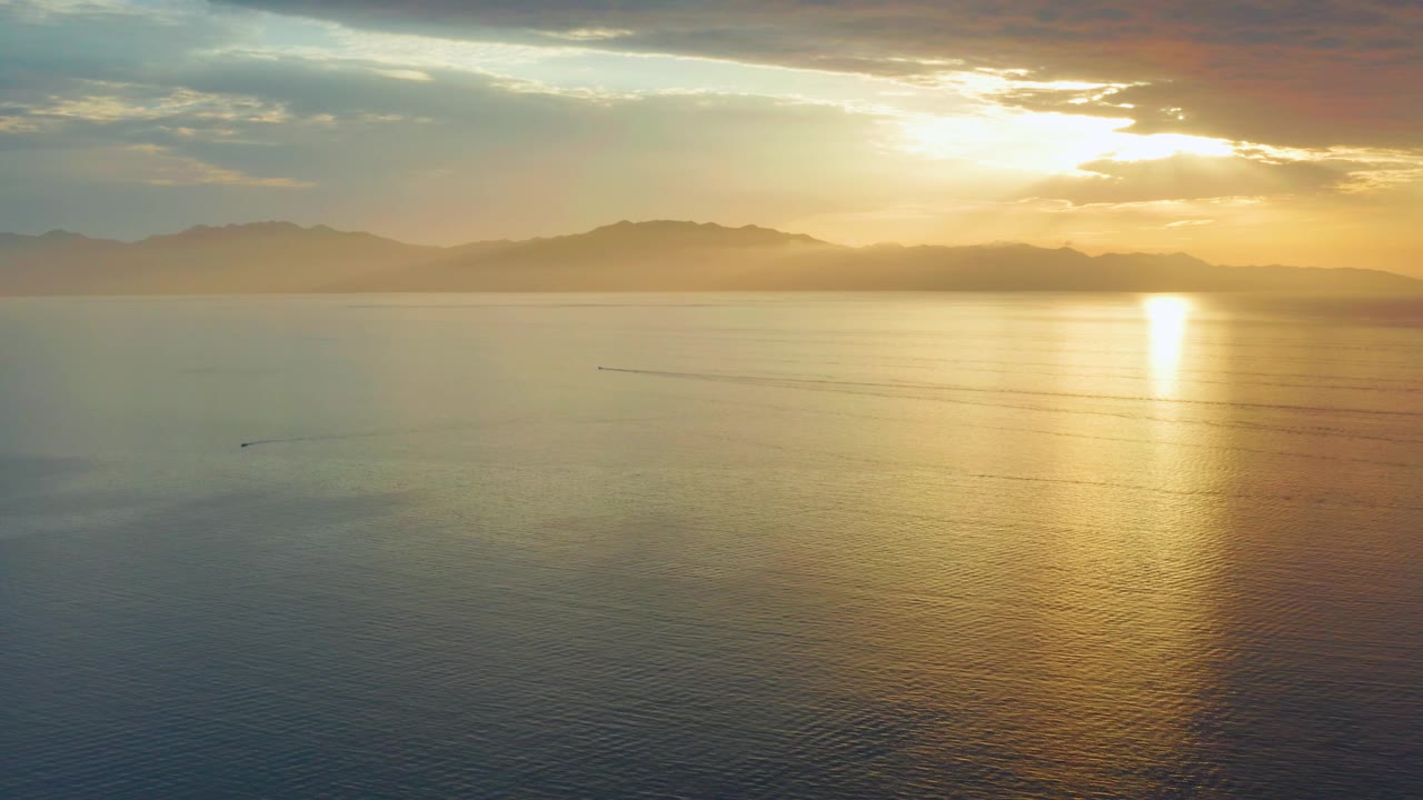 At 0:02, the frame shows a broad aerial view over the ocean in El Sargento La Ventana, with hazy mountains across the horizon, a gold reflection on the water, and no visible people.