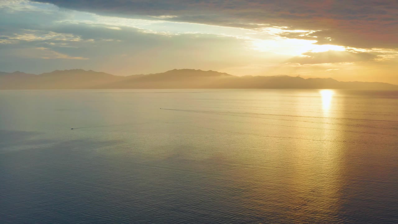 A wide aerial frame over El Sargento La Ventana, Mexico, shows blue-green water in the foreground, orange sunset light at right, mountain silhouettes on the horizon, and no visible people.