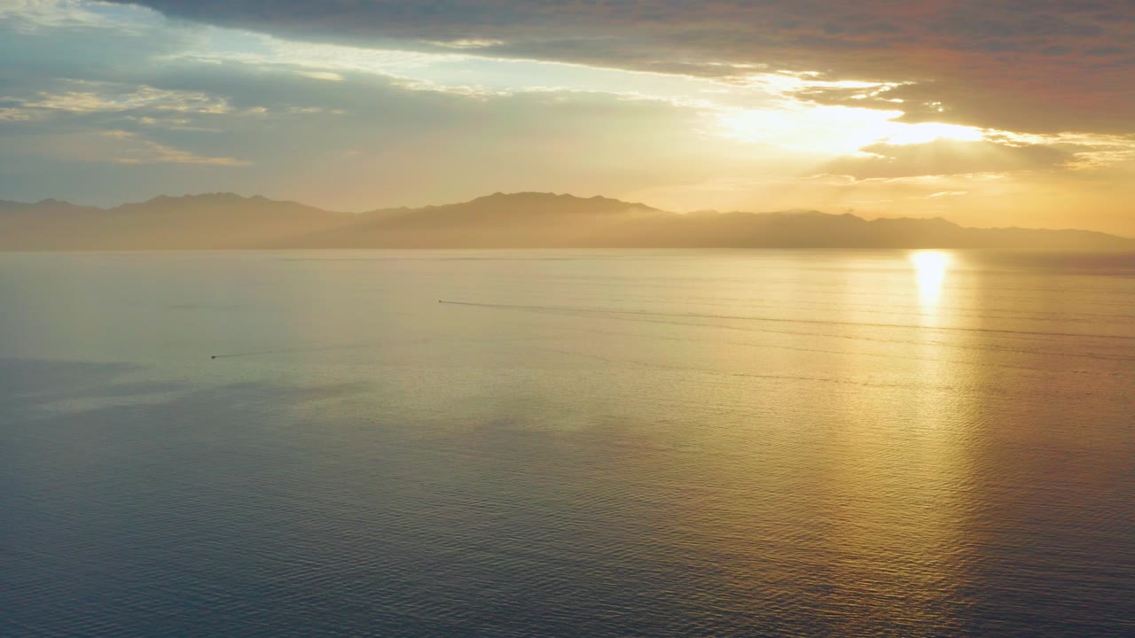 At 0:00 in El Sargento La Ventana, Mexico, golden sunlight reflects across calm ocean water below distant mountain silhouettes and layered clouds, with no visible people.