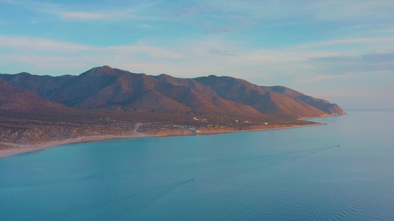 Opening aerial view over El Sargento La Ventana, Mexico, with blue water below, a curved shoreline, brown mountain ridges, soft clouds, and no visible people.
