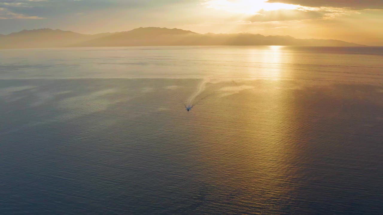 Aerial sunset frame from El Sargento La Ventana, Mexico, with the small boat slightly farther across reflective water, a bright gold path from the sun, hazy mountains, and no visible people.