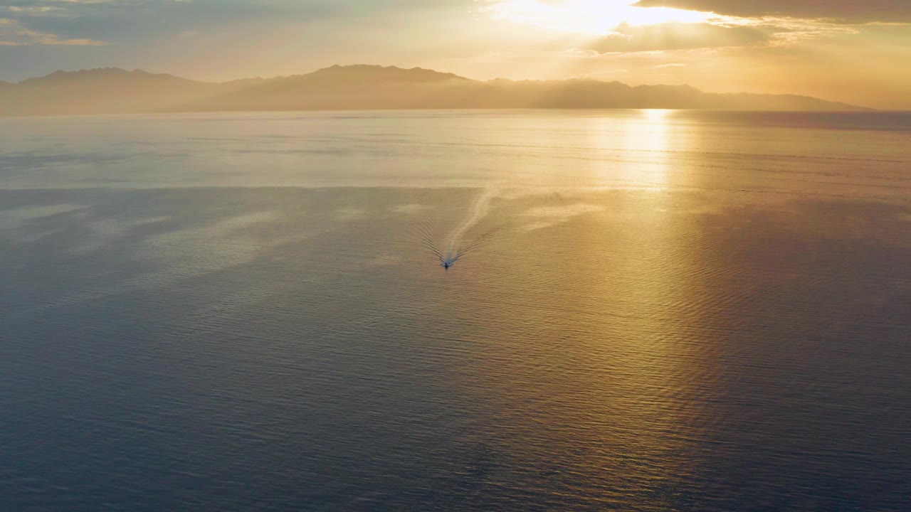 El Sargento La Ventana, Mexico appears as distant land under warm clouds while a small boat moves through the mid-lower water and the sun reflection widens across the sea, with no visible people.