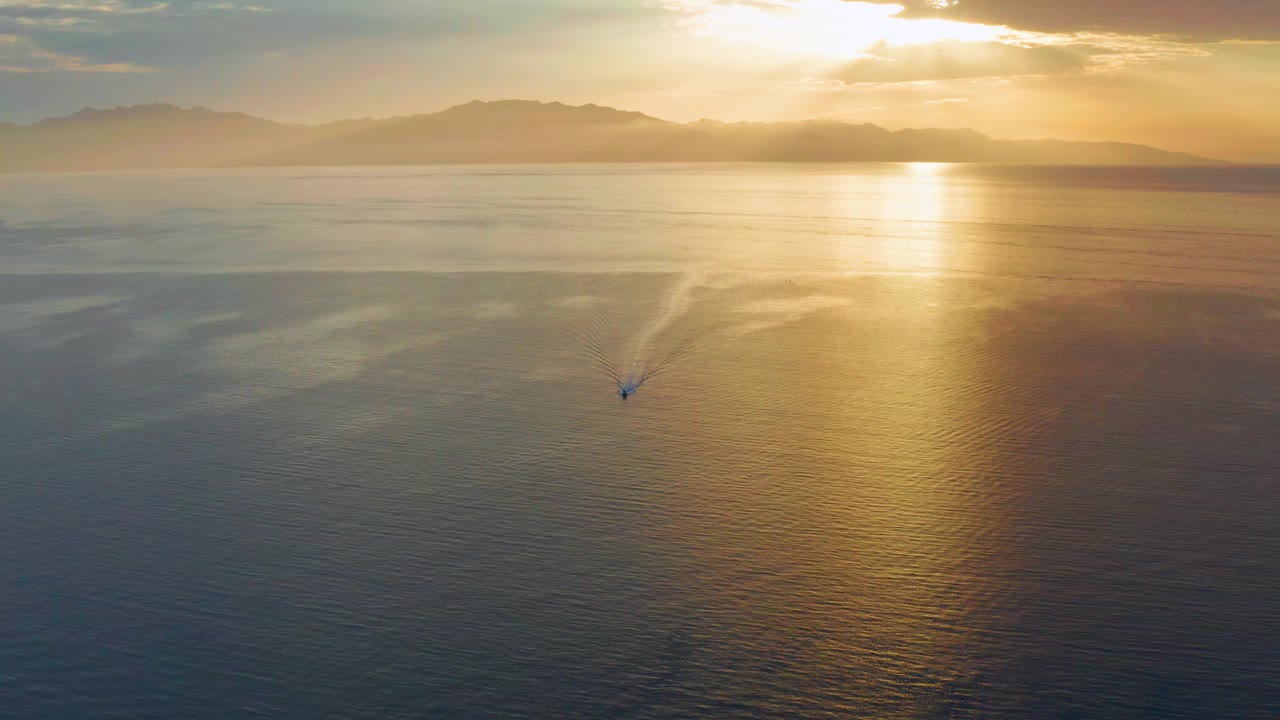 At 0:01, the El Sargento La Ventana aerial view keeps the sun high at upper right, with its reflection stretching down the sea and a small wake shifting slightly below center, with no visible people.