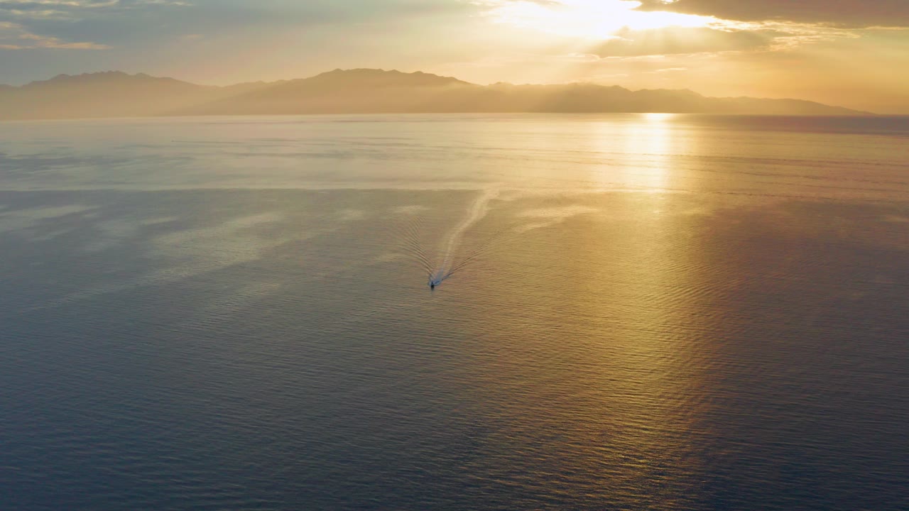 Aerial view over the sea at El Sargento La Ventana, Mexico, with golden morning light reflecting on the water, distant hills on the horizon, and a tiny subject leaving a wake near the lower center.