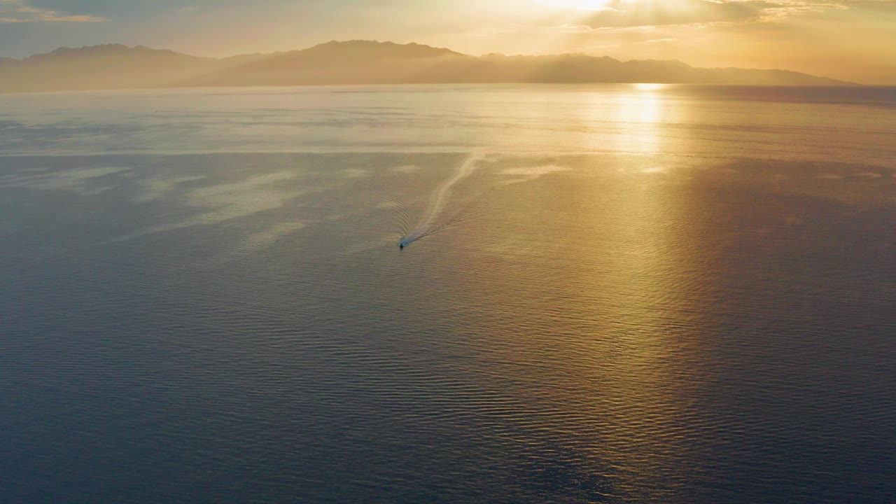 Aerial view in El Sargento La Ventana, Mexico, with a lone small boat crossing rippled open water under a broad gold sunset reflection and no visible people.