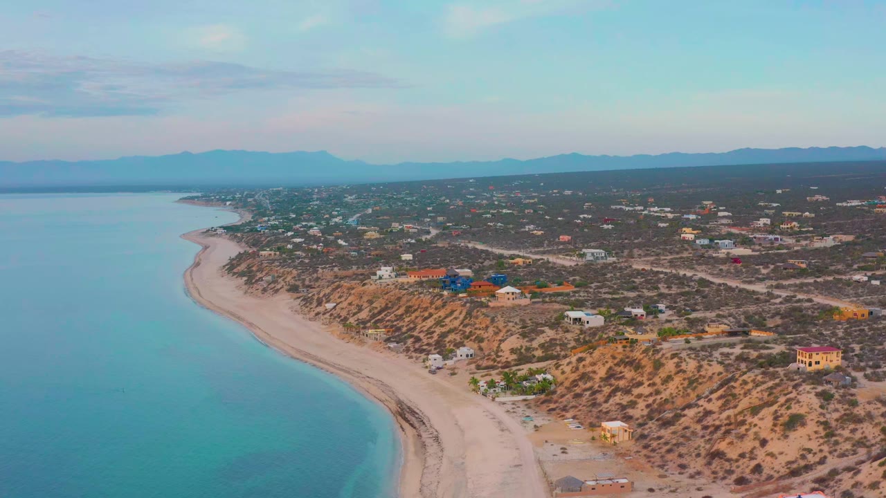 High view of the El Sargento La Ventana shore, where blue-green water meets a long sand strip, with desert lots, low houses, dirt roads, mountains, and no visible people.