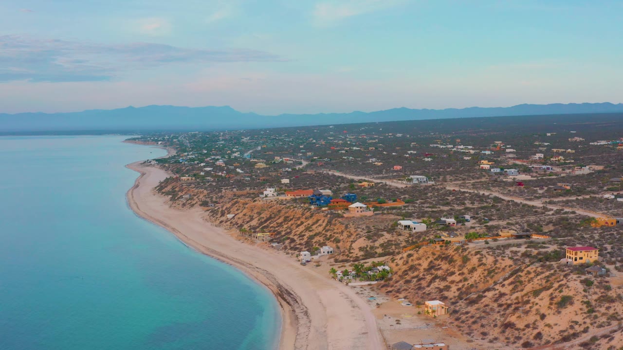 Drone-style view over El Sargento La Ventana, Mexico, showing a turquoise bay at left, pale shoreline below, clustered houses across scrub terrain, a level mountain horizon, and no visible people.