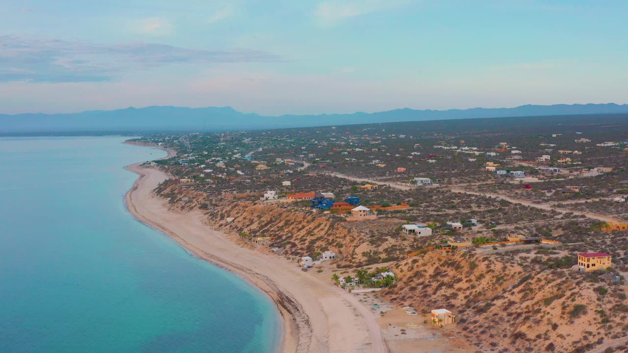 Wide aerial frame of the El Sargento La Ventana coastline, with calm sea filling the left side, a narrow sand beach below, scattered buildings inland, mountains in haze, and no visible people.