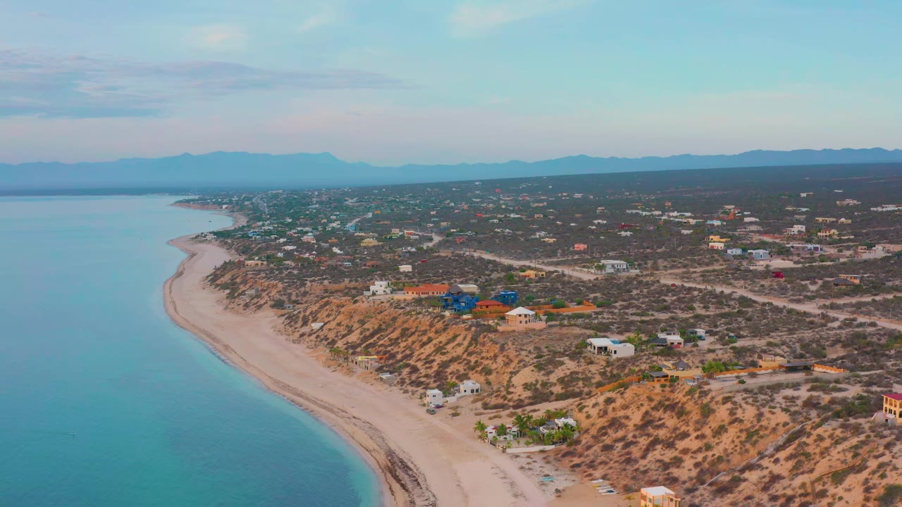 Aerial coast view with calm water, sandy shoreline, desert vegetation, and scattered town buildings.
