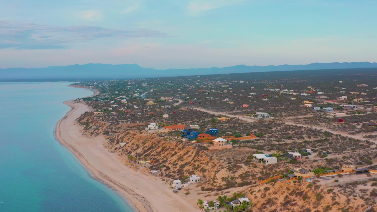 Aerial frame over El Sargento La Ventana, Mexico showing calm water at left, a pale curved beach, scattered cliffside homes, dry roads, distant mountains, and no visible people.