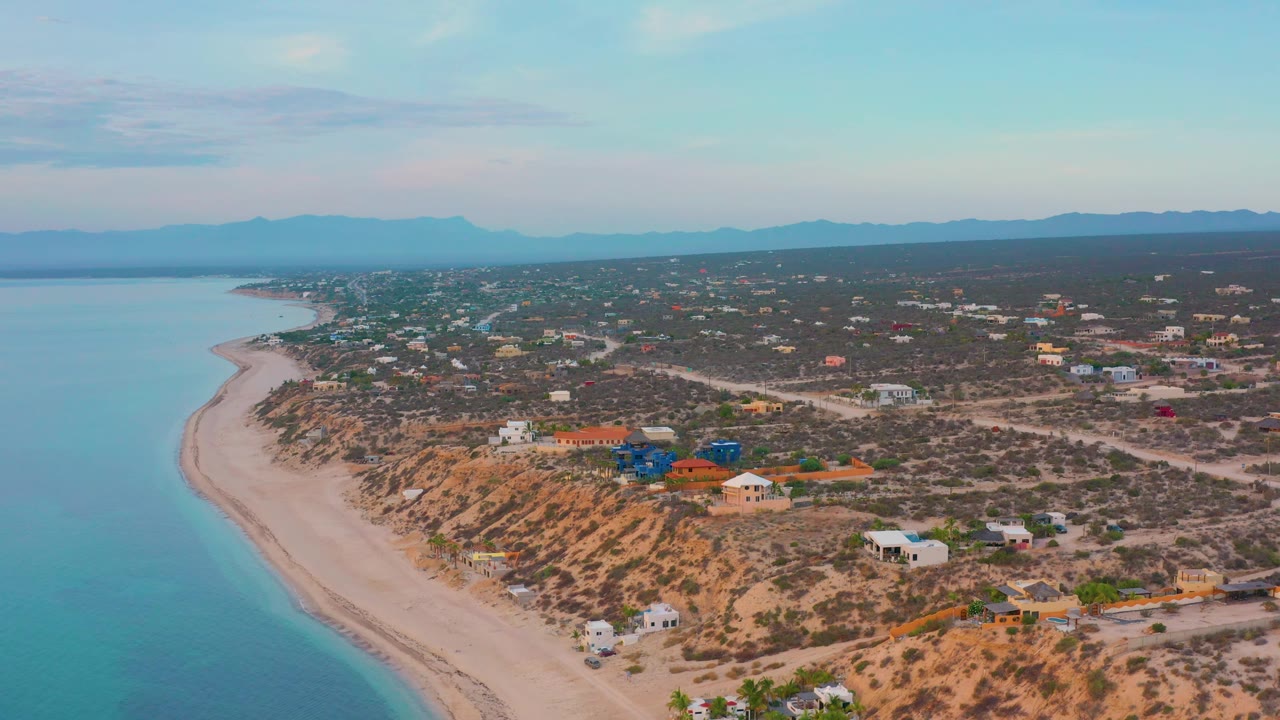 Aerial view of El Sargento La Ventana, Mexico with calm blue sea along the left edge, a pale beach below the cliff, clustered homes on dry terrain, and no visible people.
