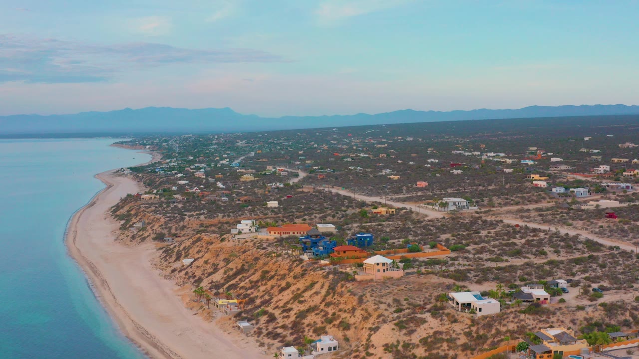 Aerial view over El Sargento La Ventana, Mexico, with a curved pale beach at left, scattered homes on scrubby hills, distant mountains, and no visible people.