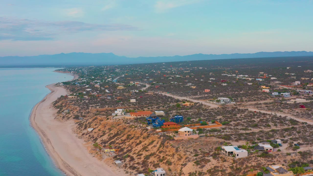 High oblique aerial over El Sargento La Ventana, Mexico, with turquoise water edging a pale beach, scattered houses on dry terrain, and no visible people.