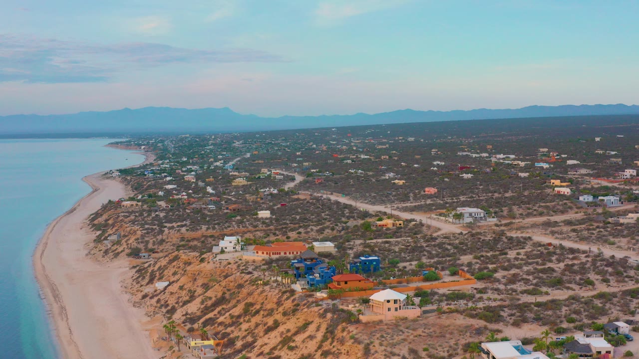 Aerial view over El Sargento La Ventana, Mexico, with blue sea and pale beach at left, scattered buildings across scrubland, distant mountains, and no visible people.