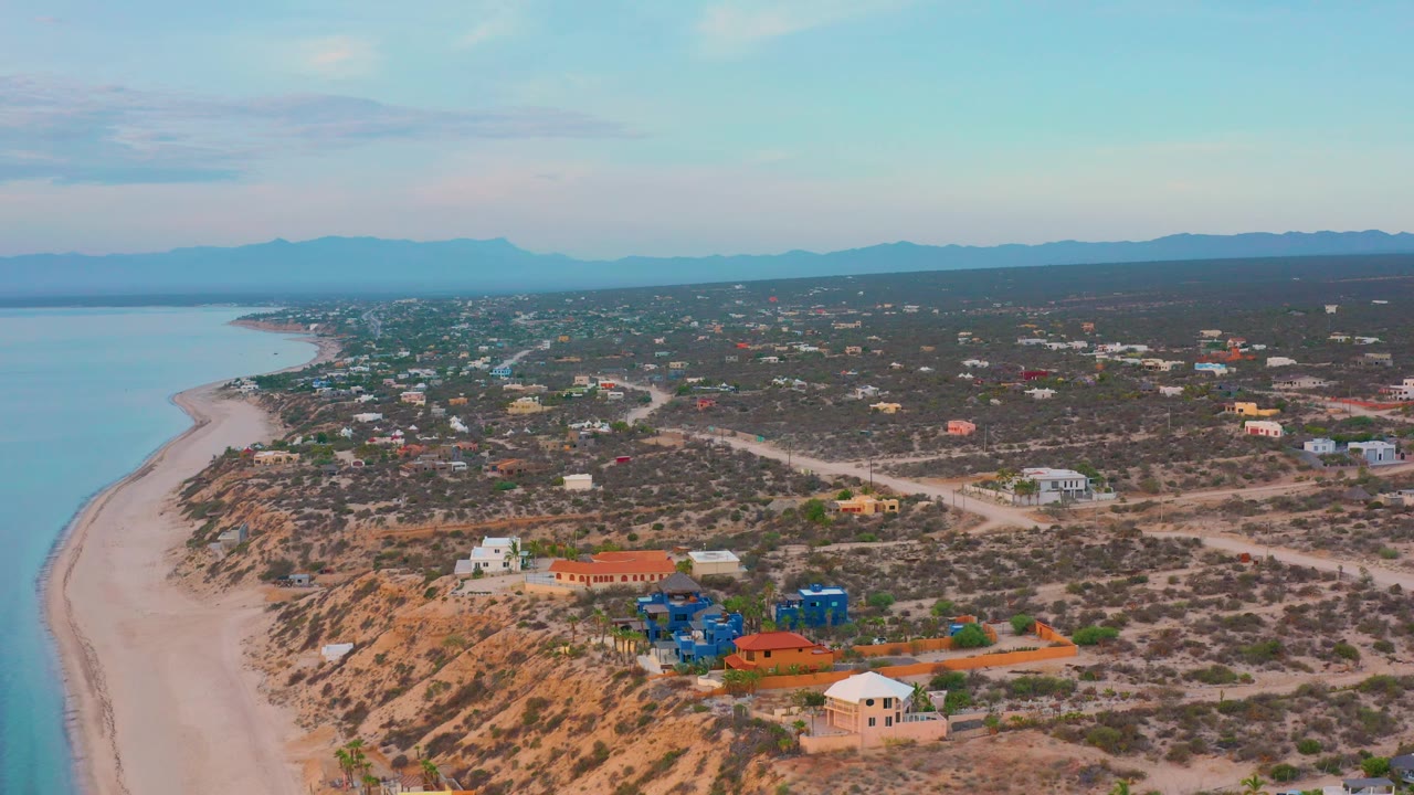 Wide aerial shoreline with scrubland, homes, roads, sea, and mountain horizon.