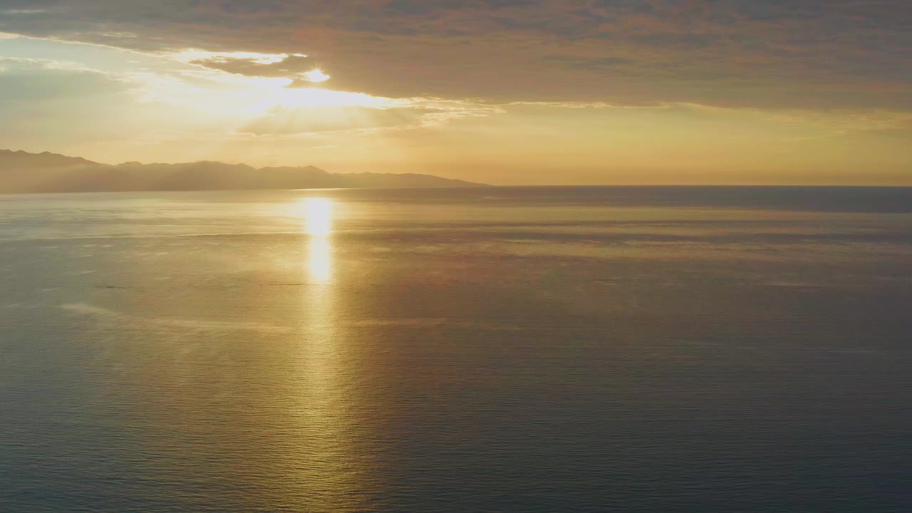 A second sunrise frame in El Sargento La Ventana, Mexico, shows the sun glow slightly stronger through the cloud break, a vertical gold path on the ocean, distant mountains, and no visible people.