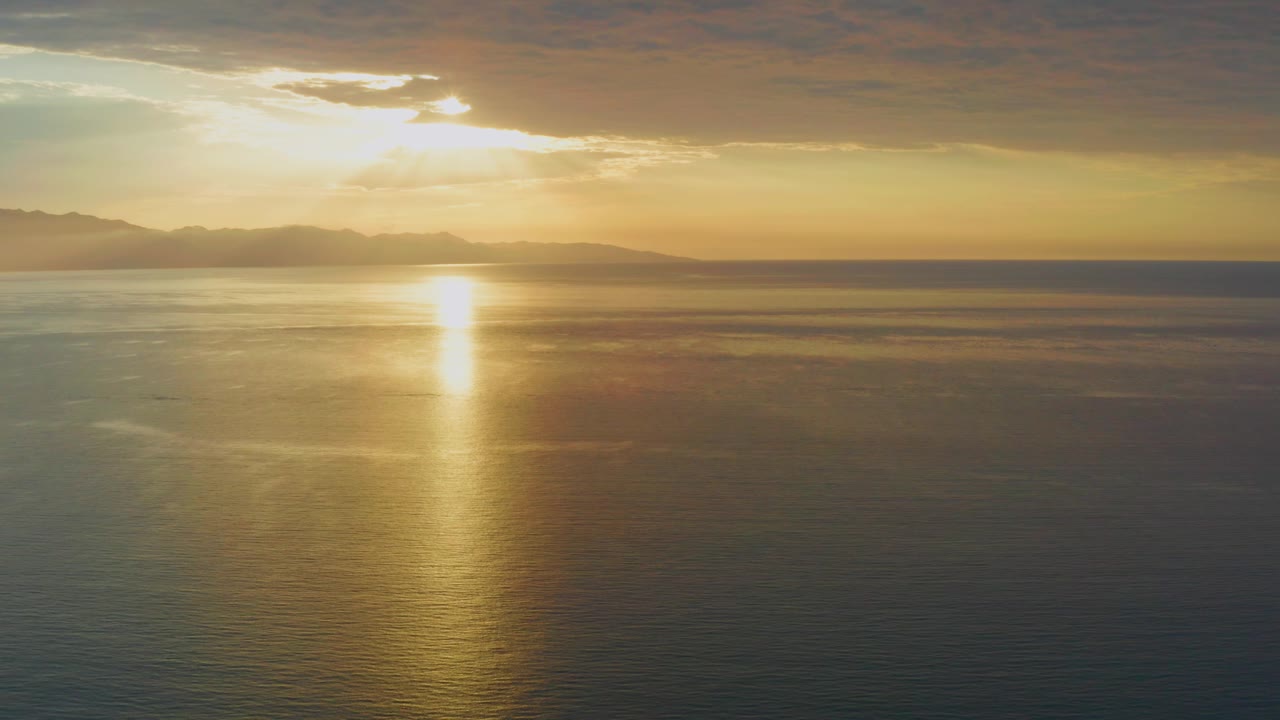 The sun sits behind broken cloud over El Sargento La Ventana, Mexico, with gold light on the ocean, layered mountain silhouettes across the horizon, and no visible people.