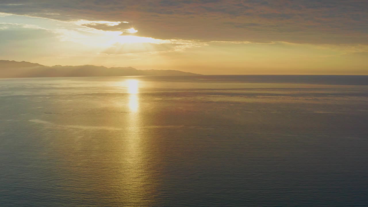 Mid-clip, the El Sargento La Ventana sunrise holds on quiet coastal water, with the bright reflection near center, subdued blue-green ocean tones, hazy mountains, and no visible people.