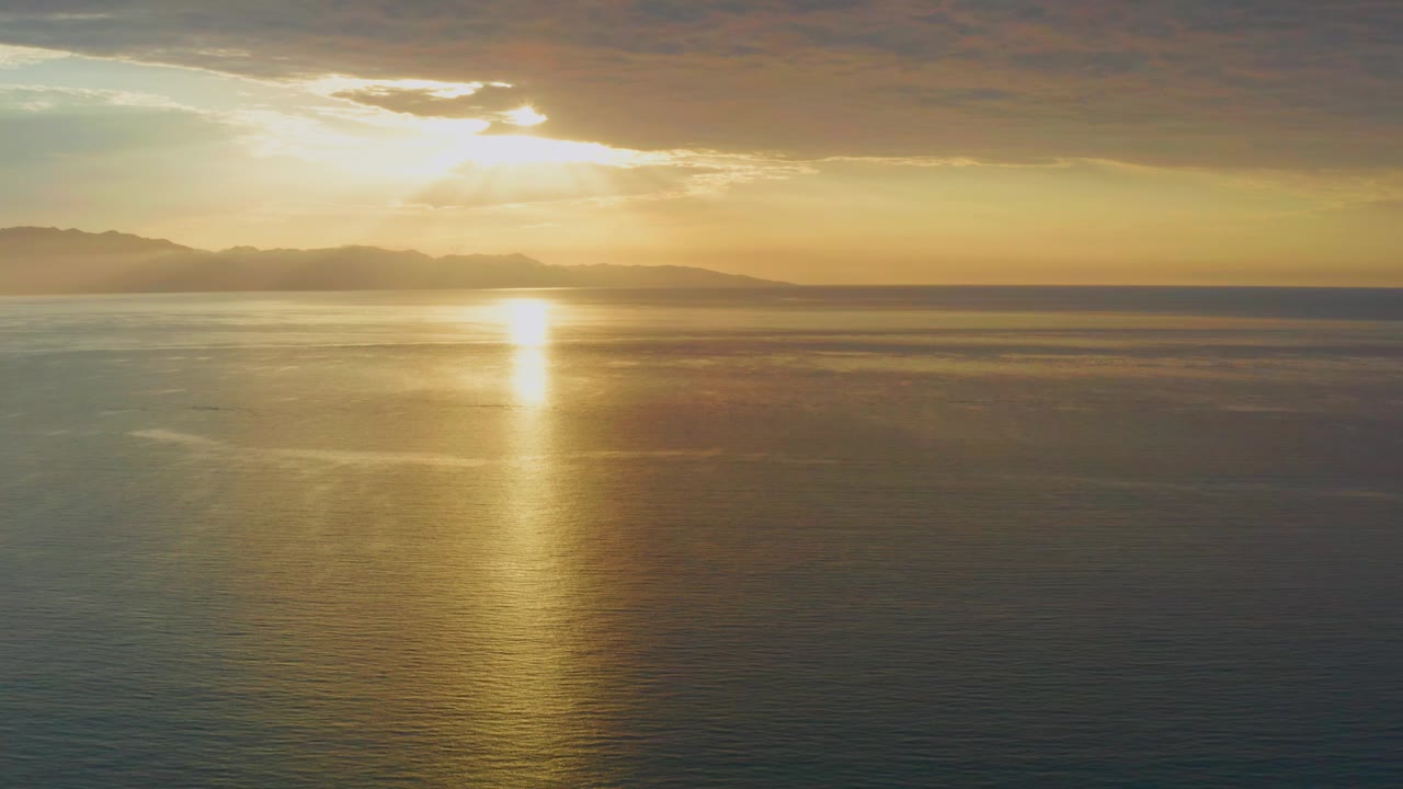 The sun sits behind a break in layered clouds above the ocean in El Sargento La Ventana, Mexico, with warm yellow light stretching across the water and no visible people.