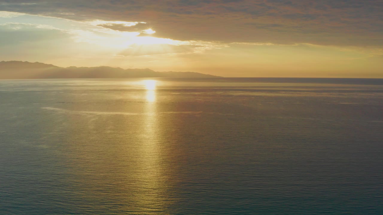 Opening frame of a low sun over the calm sea near El Sargento La Ventana, Mexico, with golden reflection on the water, a dark cloud band above, and no visible people.