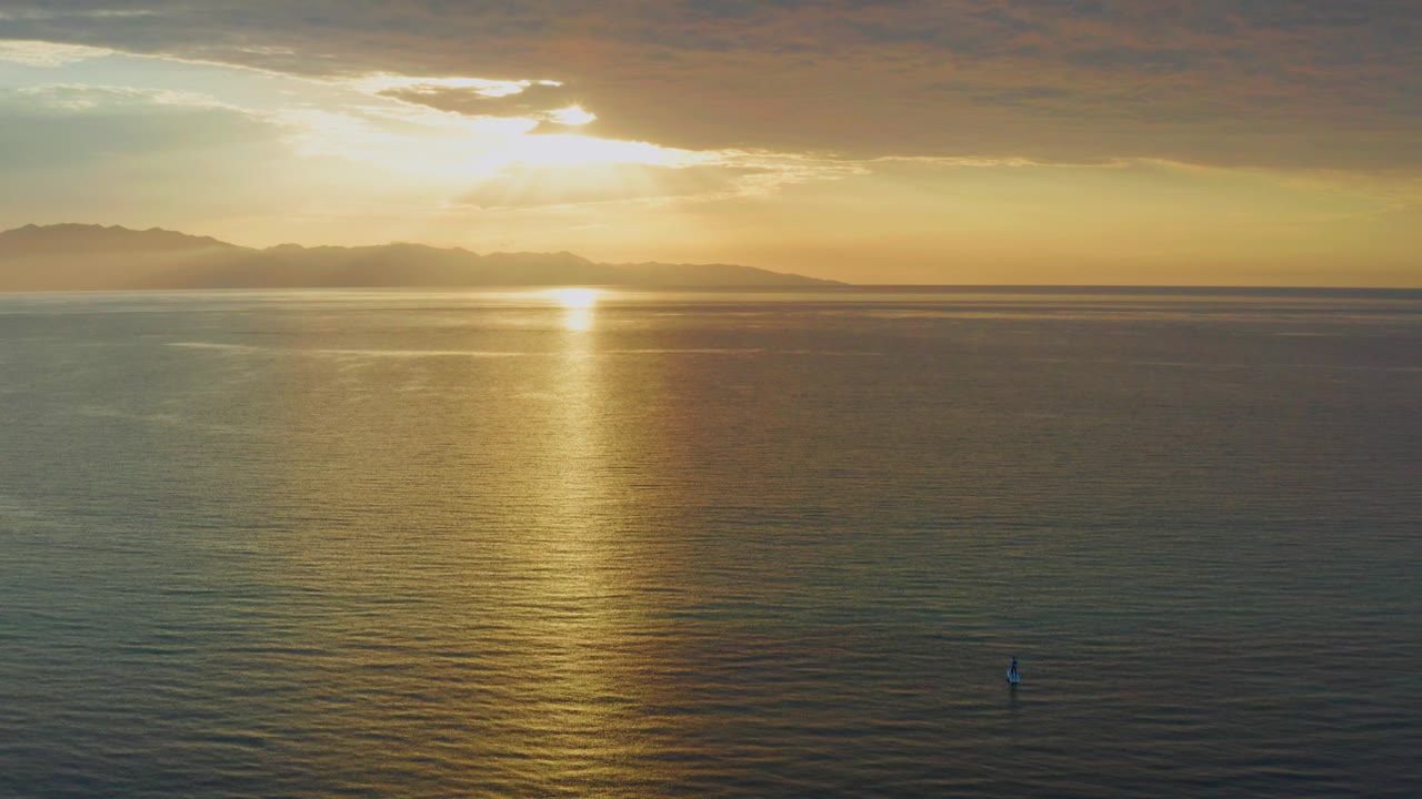 A wide aerial-looking view over El Sargento La Ventana, Mexico shows the sun glow partly under clouds, warm reflection on the sea, distant mountain silhouettes, and a very small visible person.