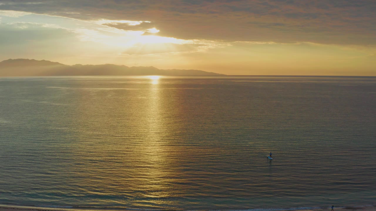Golden sunset over the ocean at El Sargento La Ventana, Mexico, with distant mountains on the horizon and a narrow strip of pale beach in the foreground.