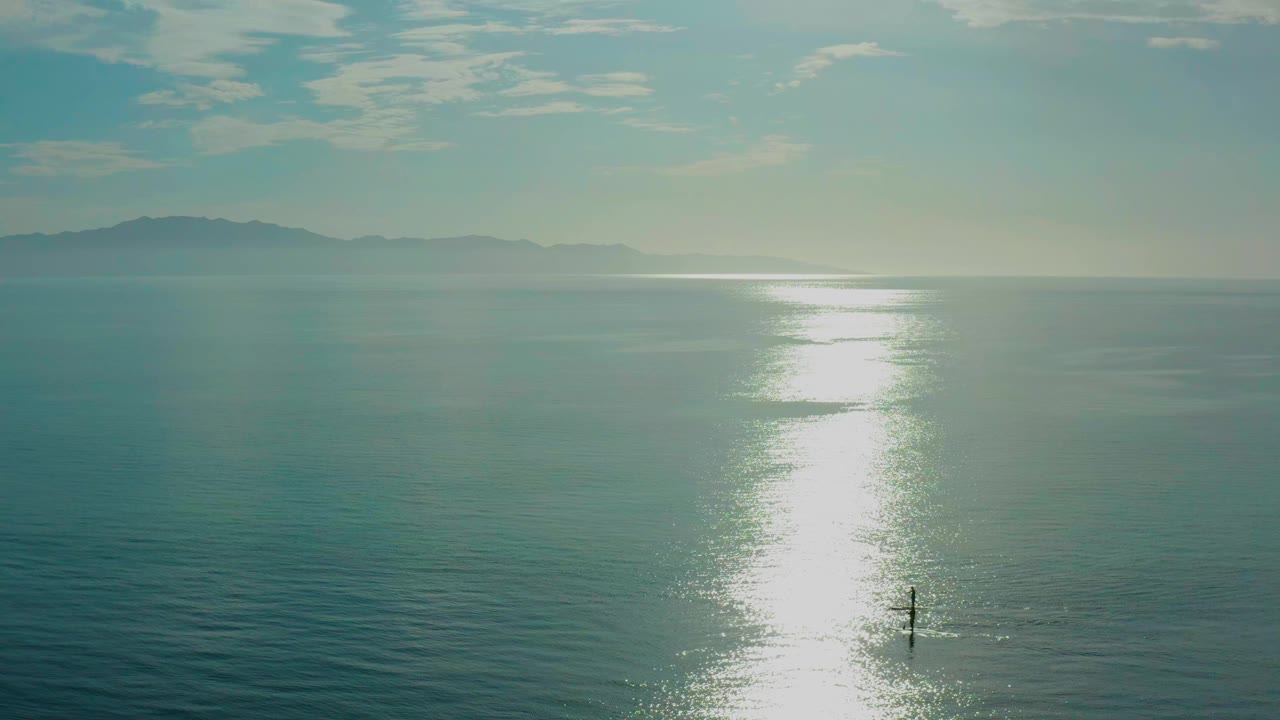 Opening frame of El Sargento La Ventana water with a bright vertical sun reflection, distant mountain horizon, and a tiny person on a sandbar near the lower center.