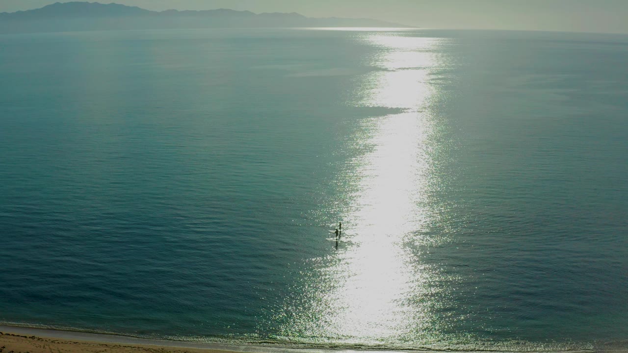 At El Sargento La Ventana, Mexico, two small paddlers sit left of a bright vertical sun reflection on teal ocean water, with a thin sandy shore at the bottom edge.