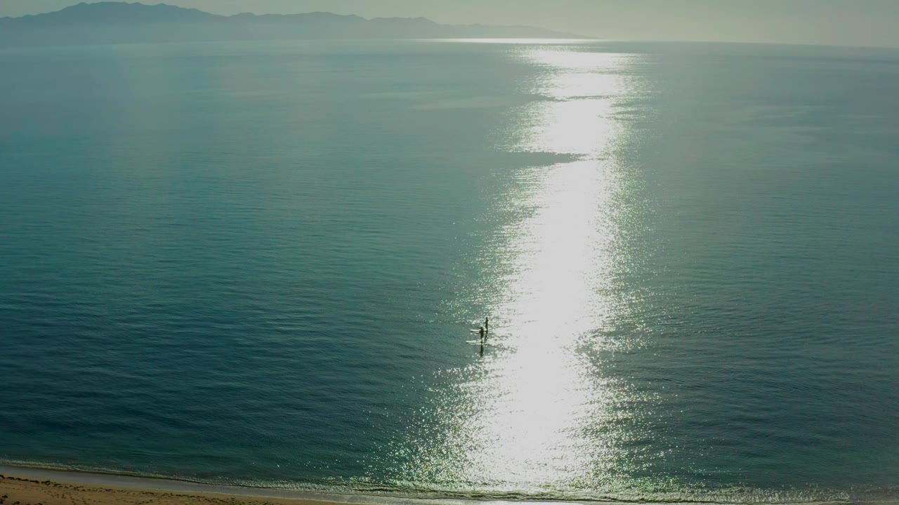 Distant paddlers beside a tall band of sun glare on calm ocean water.