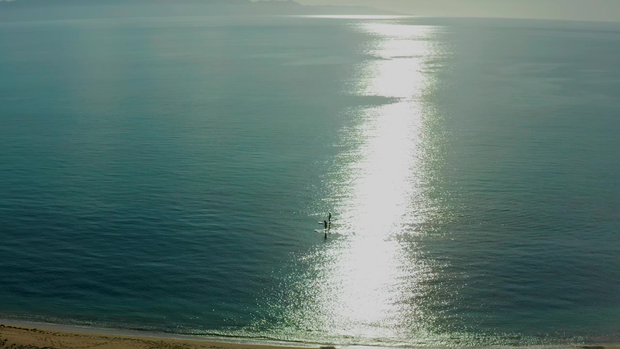 At El Sargento La Ventana, Mexico, two tiny paddlers remain near the reflected sun strip, with darker teal water to the right and pale haze along the horizon.