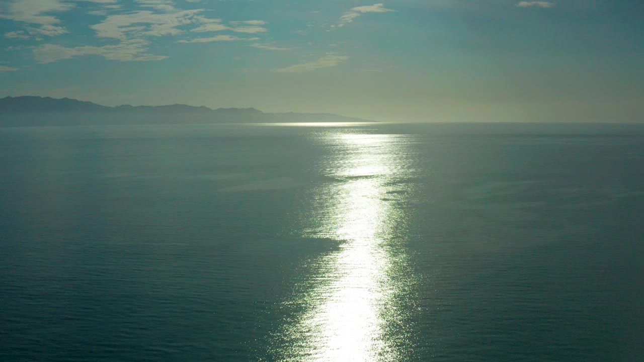 Wide view of sun glare stretching across calm water near El Sargento La Ventana, Mexico, with hazy distant land and no visible people.