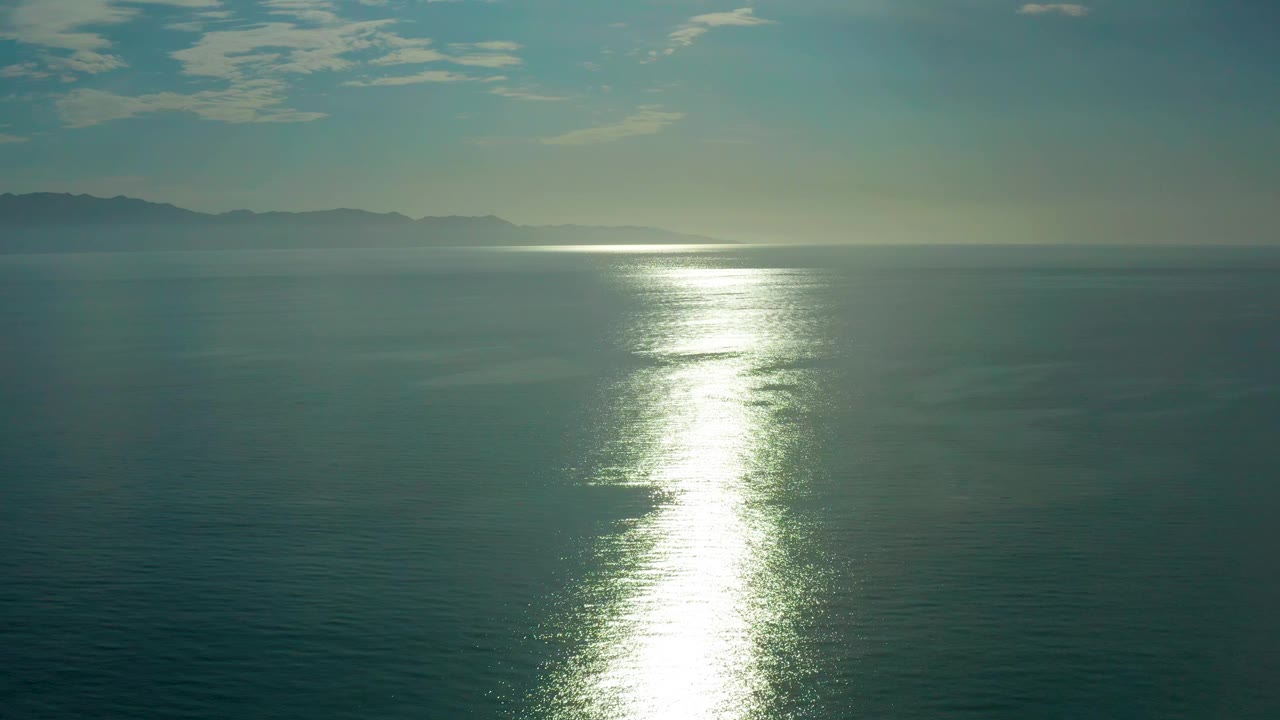 A bright strip of sunlight breaks across the water while low mountain shapes sit on the hazy horizon near El Sargento La Ventana, Mexico, with no visible people.