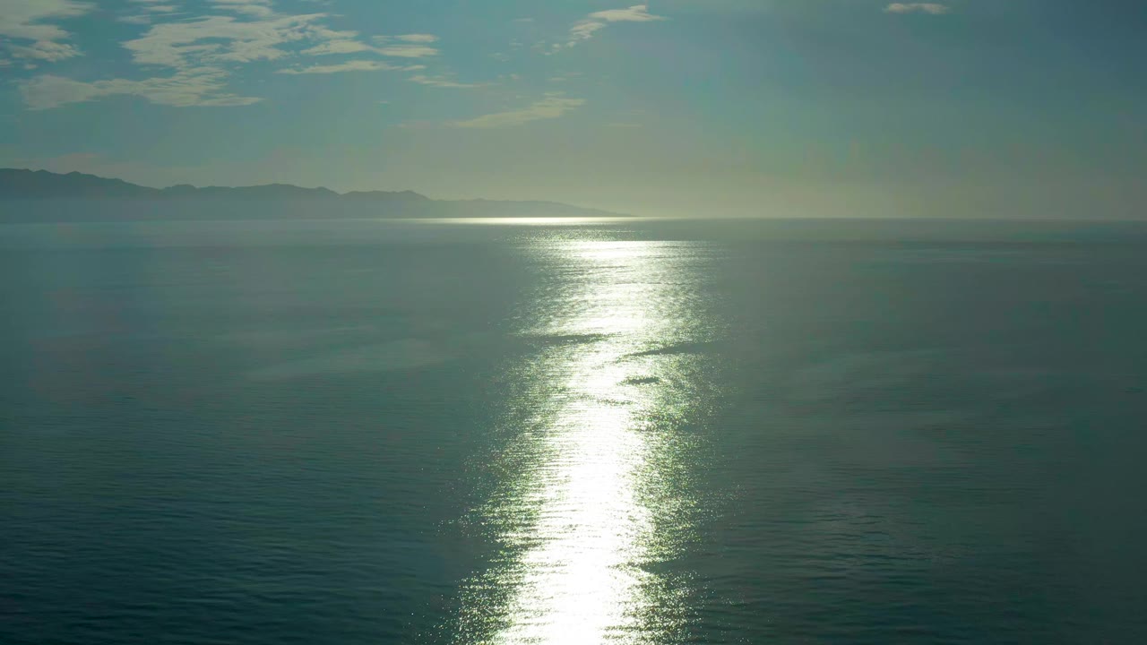 Elevated ocean view near El Sargento La Ventana, Mexico, with the sun glare centered over rippled water, low land visible in haze, small clouds above, and no visible people.