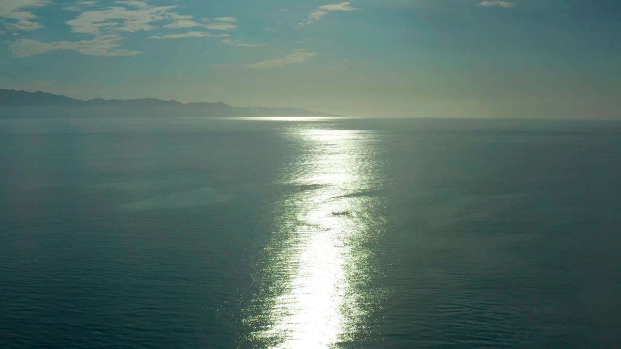 Open sea near El Sargento La Ventana, Mexico, with the bright reflection shifted low in frame, hazy land across the horizon, thin white clouds, and no visible people.