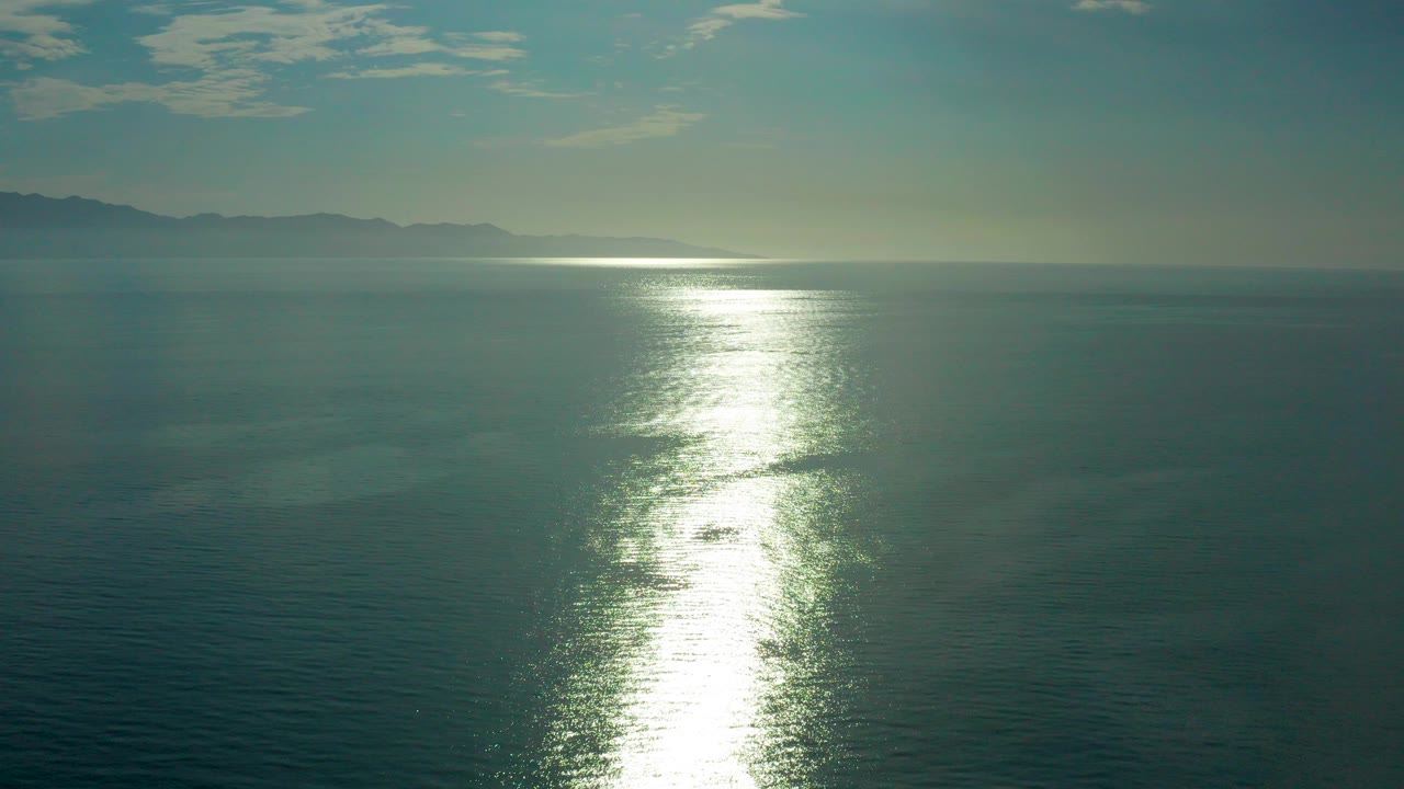 Opening frame of calm blue-green ocean in El Sargento La Ventana, Mexico, with a bright vertical sun reflection, hazy mountains on the left horizon, scattered clouds, and no visible people.
