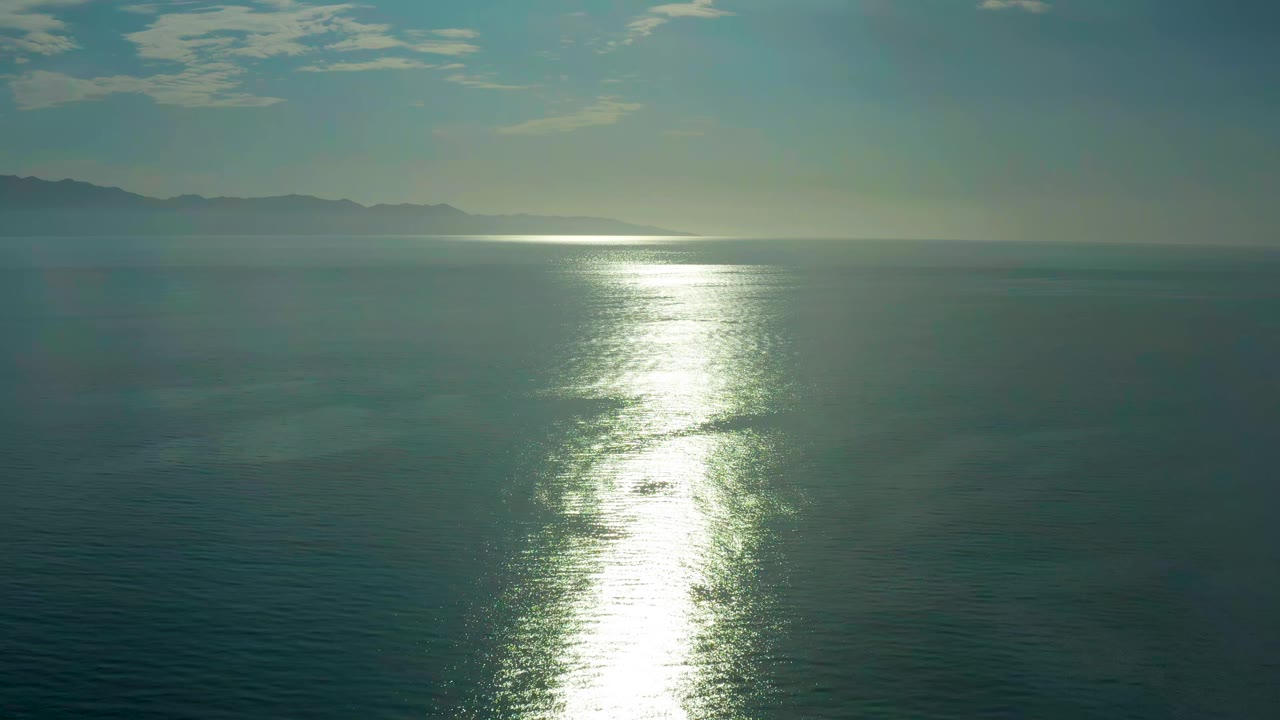 A second view of the El Sargento La Ventana ocean shows the sun path centered lower in frame, faint ripples across the water, distant land at left, and no visible people.