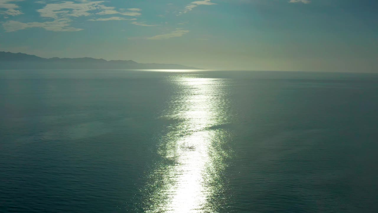 The ocean remains open and empty, with a white sun reflection stretching toward the camera, a hazy horizon, low mountain shapes to the left, and no visible people.
