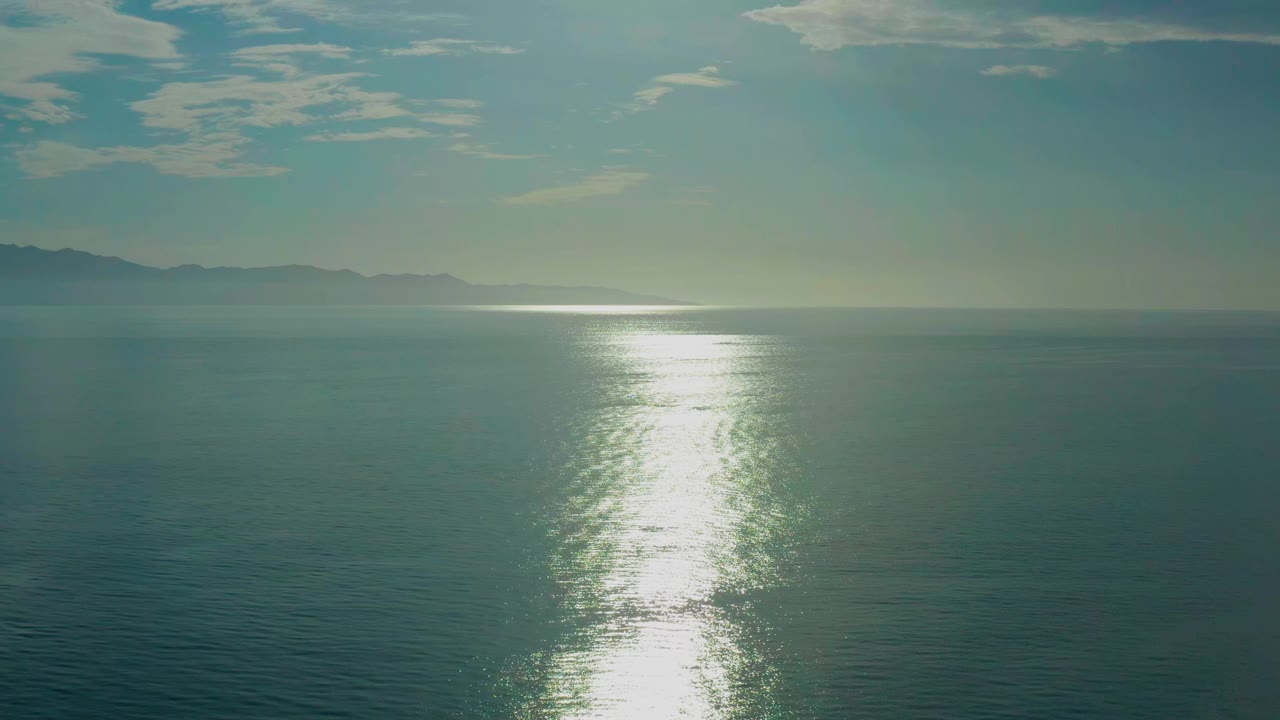 Calm blue-green water fills the foreground in El Sargento La Ventana, Mexico, while the sun reflection runs vertically toward the camera and low mountains sit along the left horizon with no visible people.