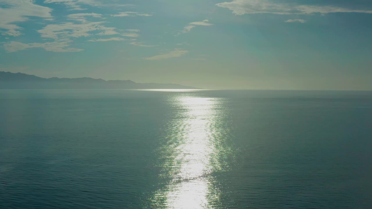 A centered band of sun glare brightens the ocean in El Sargento La Ventana, Mexico, with muted teal water below, pale sky above, distant land to the left, and no visible people.