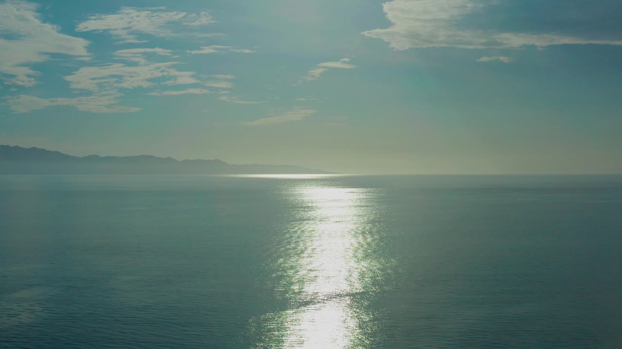 Opening frame of blue green sea near El Sargento La Ventana, Mexico, with a bright vertical sun reflection, distant left coastline, pale clouds, and no visible people.
