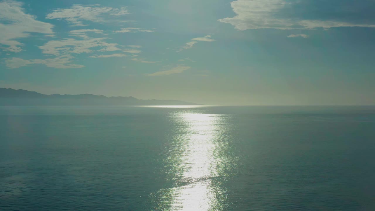 Aquamarine water fills the frame at El Sargento La Ventana, Mexico, with the sun path shimmering near center, a low distant horizon, cloud cover above, and no visible people.