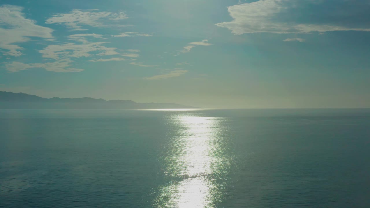 Mid-clip frame of calm ocean at El Sargento La Ventana, Mexico, with a bright vertical reflection on the lower water, hazy horizon, soft clouds, and no visible people.