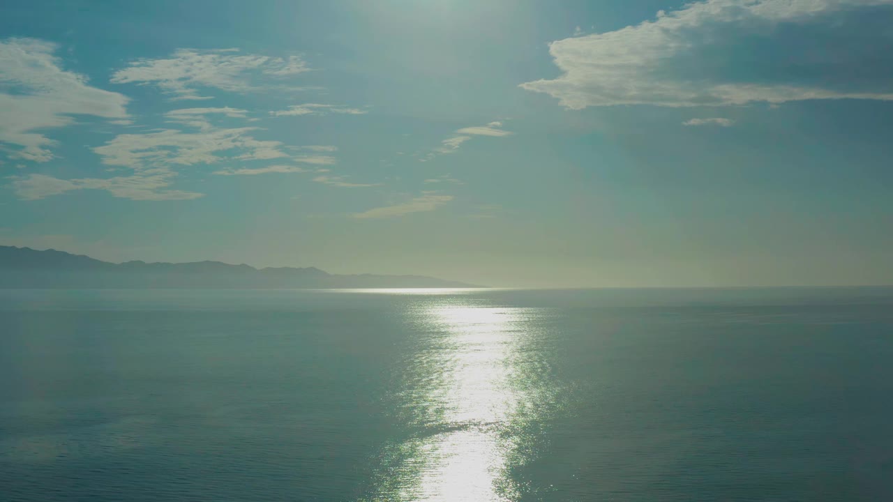 At 0:01, the vertical band of reflected sunlight sits slightly left of center on the ocean near El Sargento La Ventana, Mexico, with cloud shapes spread across the upper sky and no visible people.